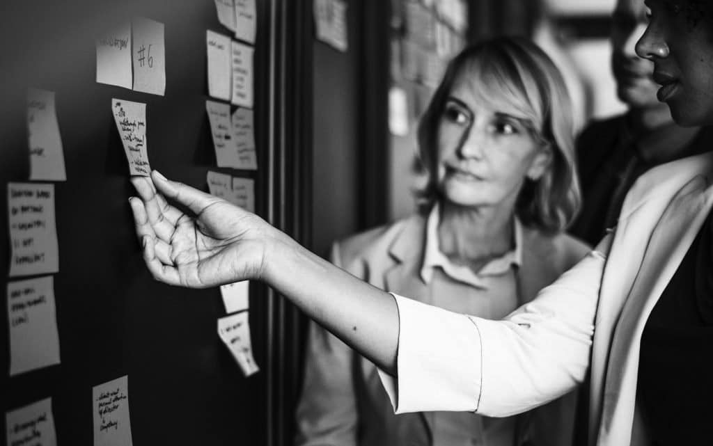 Black and white image of two women looking at board of post-it notes. One of them is reaching out and touching one to read it.