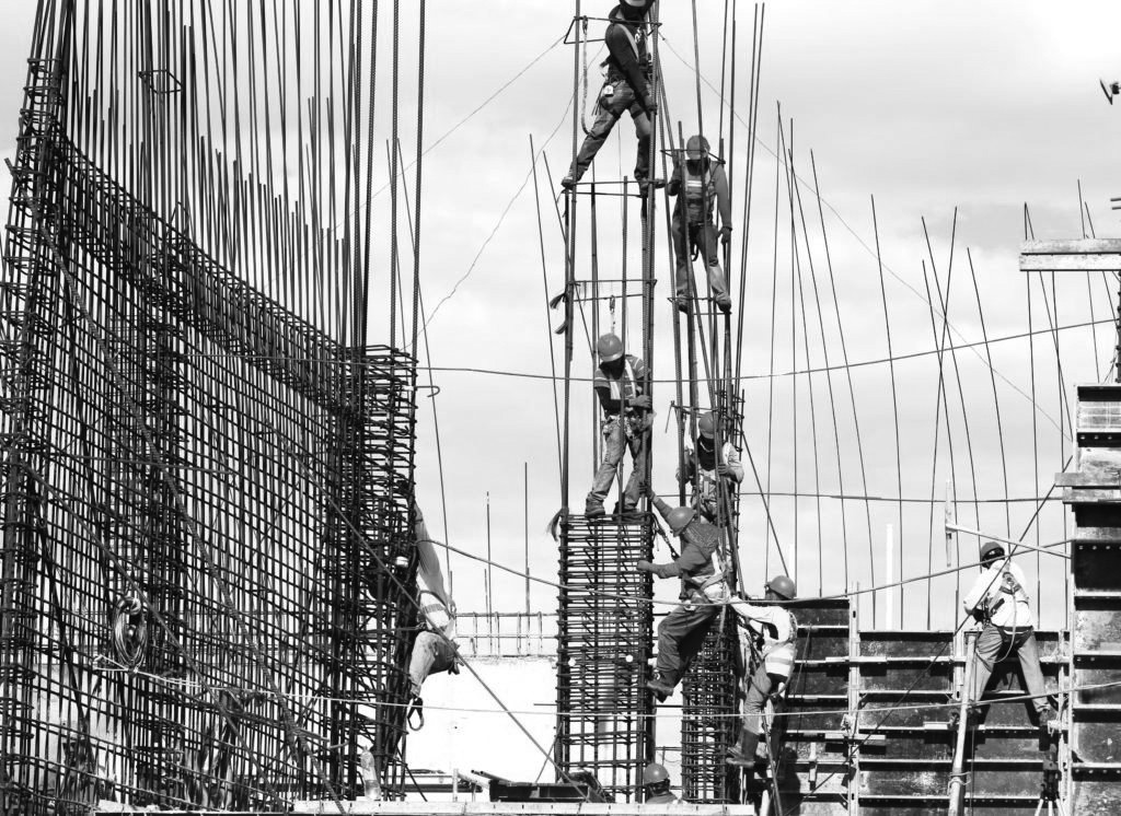 Black and white image of several people climbing fences of metal wire wearing construction clothing and helmets whilst helping each other up